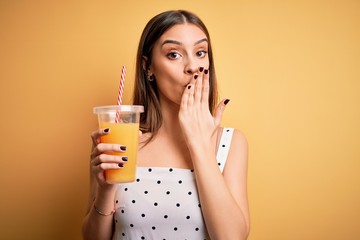 Young beautiful brunette woman drinking healthy orange juice over yellow background cover mouth with hand shocked with shame for mistake, expression of fear, scared in silence, secret concept