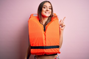 Young beautiful brunette woman wearing orange lifejacket over isolated pink background with a big...