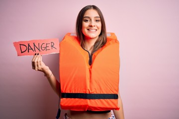 Young beautiful brunette woman wearing lifejacket holding paper with danger message with a happy...