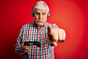 Senior handsome hoary gamer man playing video game using joystick and headphones pointing with...