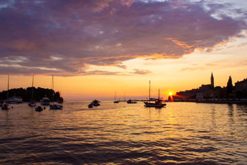 Fototapeta na wymiar Adriatic sea with some boats sailing and the view of the croatian houses of Rovinj, Croatia, and the bell tower of the Church of St Euphemia at the background, during the sunset