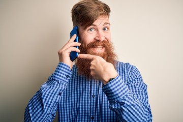 Redhead Irish man with beard having a conversation talking on smartphone over isolated background very happy pointing with hand and finger