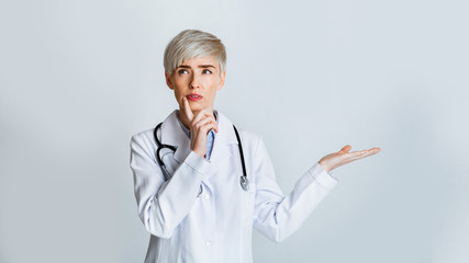 Pensive woman doctor press finger to chin and held up palm for empty space
