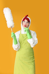 Thoughtful housewife in protective costume and with dust brush on color background