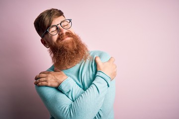 Handsome Irish redhead man with beard wearing glasses over pink isolated background Hugging oneself...