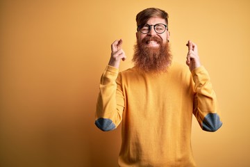 Naklejka premium Handsome Irish redhead man with beard wearing glasses over yellow isolated background gesturing finger crossed smiling with hope and eyes closed. Luck and superstitious concept.