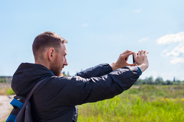 Bearded man in black raincoat on a blue sea landscape background with smartphone in hands takes seascape photography. Portrait of running young hipster guy with cell phone on nature, lifestyle photo.