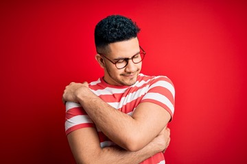Young handsome man wearing casual striped t-shirt and glasses over isolated red background Hugging...