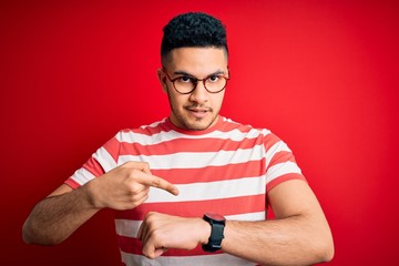 Young handsome man wearing casual striped t-shirt and glasses over isolated red background In hurry...