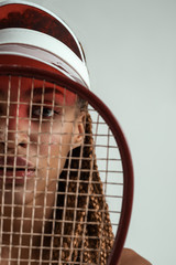 Winner. Close up portrait of beautiful young african woman holding tennis racket in front of her face and looking at camera while standing against grey background