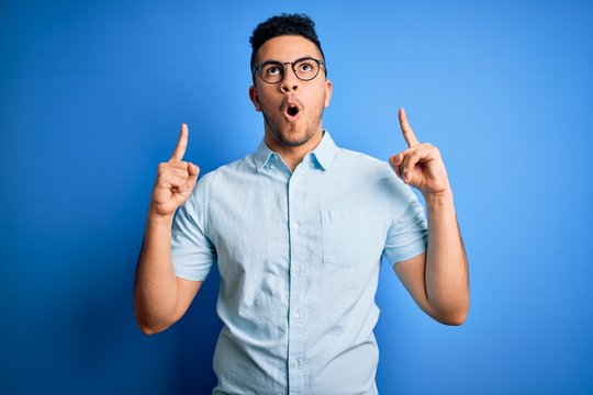 Young handsome man wearing casual summer shirt and glasses over isolated blue background amazed and surprised looking up and pointing with fingers and raised arms.