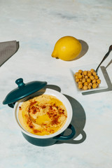 dish of hummus with olive oil and paprika. Next to it there is a lemon and a plate of chickpeas, on a white wooden board