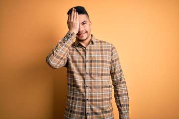 Fototapeta premium Young handsome man wearing casual shirt standing over isolated yellow background covering one eye with hand, confident smile on face and surprise emotion.