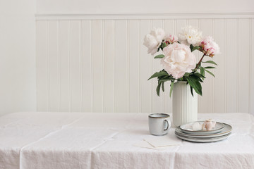 Styled stock photo. Feminine wedding or birthday table composition with floral bouquet. Pink peonies flowers, cup of coffee, plates and silk ribbons. White wooden wall background. Rustic interior.