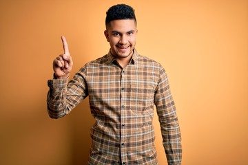 Young handsome man wearing casual shirt standing over isolated yellow background pointing finger up with successful idea. Exited and happy. Number one.