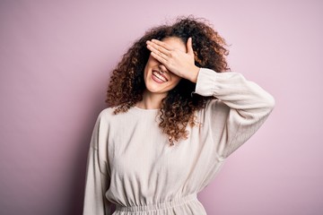 Beautiful woman with curly hair and piercing wearing casual sweater over pink background smiling and laughing with hand on face covering eyes for surprise. Blind concept.