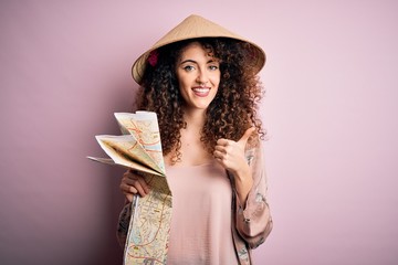 Young beautiful tourist woman with curly hair and piercing wearing asian hat holding city map happy with big smile doing ok sign, thumb up with fingers, excellent sign