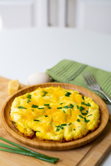 Fluffy omelette from whipped eggs with cheddar cheese and green onion scallions sprinkled on it in a wooden plate. It is surrounded by scallions, an egg, basil leaves, cheese, a green napkin, a fork