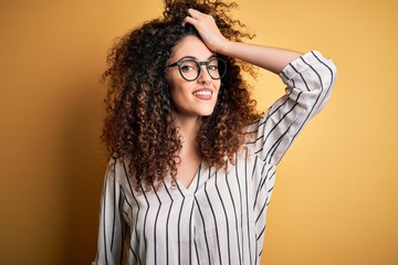 Young beautiful woman with curly hair and piercing wearing striped shirt and glasses smiling confident touching hair with hand up gesture, posing attractive and fashionable