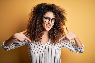 Young beautiful woman with curly hair and piercing wearing striped shirt and glasses looking confident with smile on face, pointing oneself with fingers proud and happy.