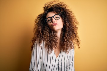 Young beautiful woman with curly hair and piercing wearing striped shirt and glasses looking at the camera blowing a kiss on air being lovely and sexy. Love expression.