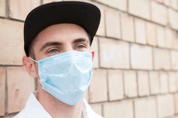 Close up of a stylish young man looking away cheerfully posing outdoors in the city, wearing protective medical mask