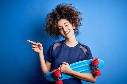 Young sporty woman with curly hair and piercing holding skate over blue background very happy pointing with hand and finger to the side