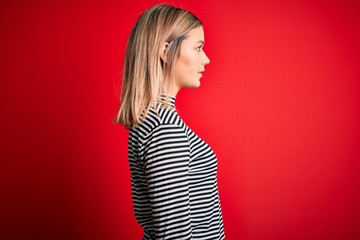 Young beautiful blonde woman wearing casual striped sweater over red isolated background looking to...