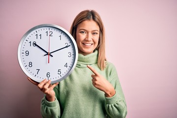 Young beautiful woman holding clock standing over isolated pink background very happy pointing with hand and finger