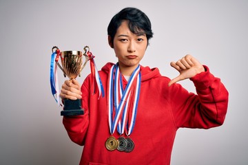Young beautiful asian girl winner holding trophy wearing medals over white background with angry...