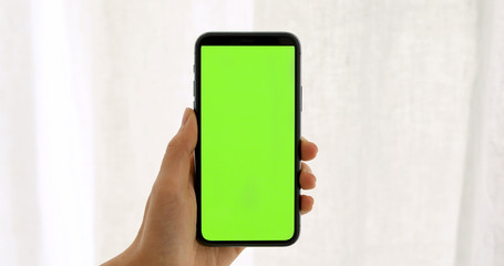 Close-up of a woman's hand holding green mock-up screen smartphone. Modern mobile phone. In the background cozy white curtain