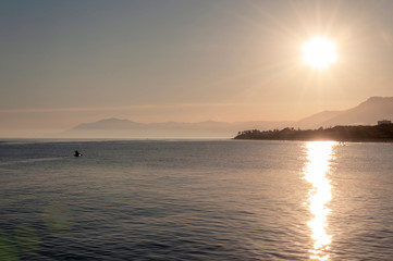 Fototapeta premium Silhouette of a man on a kayak against the backdrop of the setting sun with a bright glare on the sea on the background of the coast with palm trees and mountains