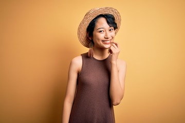 Young beautiful asian girl wearing casual t-shirt and hat over isolated yellow background looking confident at the camera with smile with crossed arms and hand raised on chin. Thinking positive.