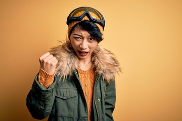 Beautiful asian skier girl wearing snow sportswear using ski goggles over yellow background angry and mad raising fist frustrated and furious while shouting with anger. Rage and aggressive concept.
