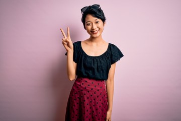 Young beautiful asian girl wearing casual dress standing over isolated pink background smiling with happy face winking at the camera doing victory sign with fingers. Number two.