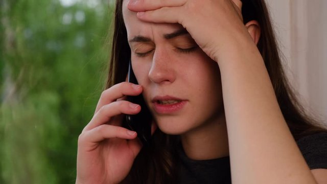 Sad unhappy woman sit on windowsill placing hand on head when hearing bad news on cellphone, tired female from too long talking on mobile phone, girl shocked of senseless and unproductive conversation