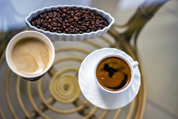 Top view of caffe latte and black coffee on glass table. Roasted coffee beans in white bowl next to two different type of coffee.  Cup of coffee to go on the glass table with turkish black coffe.