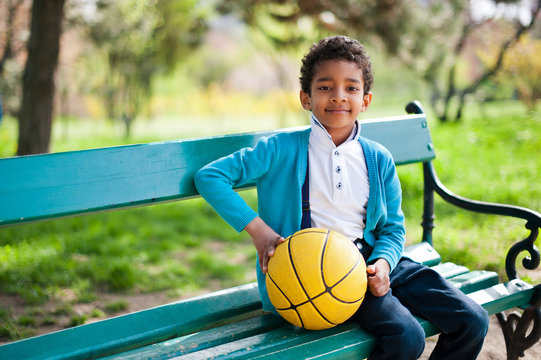 Handsome little black boy siting on a bench with a yellow ball in the park in summertime.
