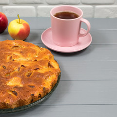 Homemade baked apple pie, in a plate, on a gray wooden table, ready to eat. A pink cup of tea. Apples