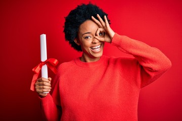 Young African American afro student woman with curly hair holding university degree diploma with happy face smiling doing ok sign with hand on eye looking through fingers