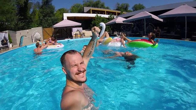 Lifestyle blogger man taking selfie video with action camera in a swimming pool. Travel vlogger films vlog from party at luxury resort. Fitted guy live streaming for social media likes. Slow motion.