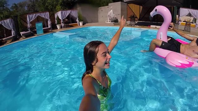 Lifestyle blogger woman taking selfie video with action camera in swimming pool. Travel vlogger films vlog from party at luxury resort. Fitted girl live streaming and relaxing with friends on vacation