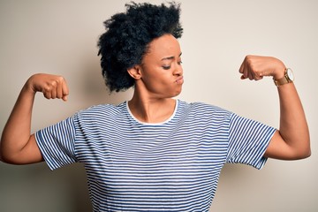 Young beautiful African American afro woman with curly hair wearing striped t-shirt showing arms muscles smiling proud. Fitness concept.