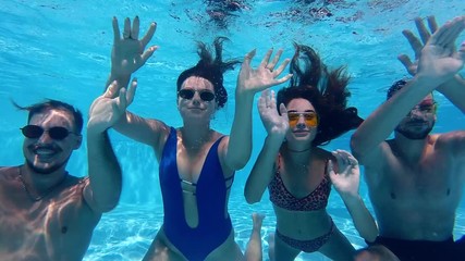 Happy friends diving under water and waving hands at action camera in swimming pool at luxury resort. Young people in swimwear having fun partying in private holiday villa. Hot girls dive underwater.