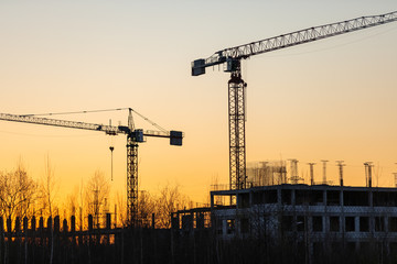 Industrial construction cranes and buildings silhouettes at a construction site on the background at sunset or dawn.