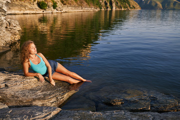 Fototapeta na wymiar Girl lying on rocky shore lowering her feet into water.