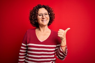 Middle age beautiful curly hair woman wearing casual striped sweater over red background smiling with happy face looking and pointing to the side with thumb up.