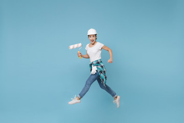 Naklejka na ściany i meble Side view funny young woman in protective helmet hardhat jump with paint roller isolated on blue background. Instruments accessories for renovation apartment room. Repair home concept. Looking camera.