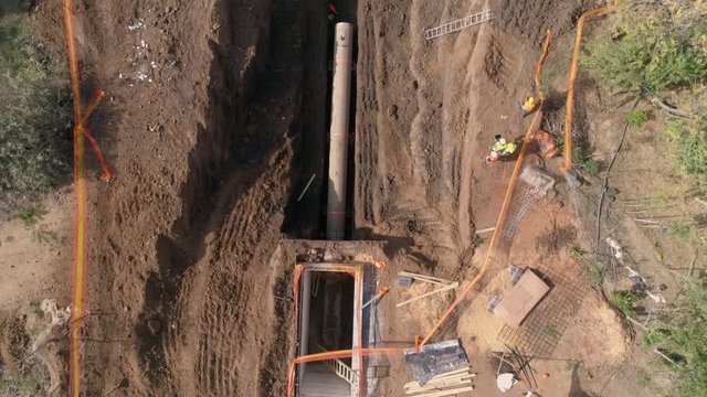 Top down aerial above construction site with workers and excavator digger working and digging on raw sand ground with huge concrete pipelines 