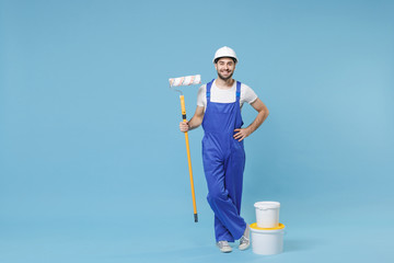 Smiling young man in coveralls protective helmet hardhat hold paint roller, bucket isolated on...